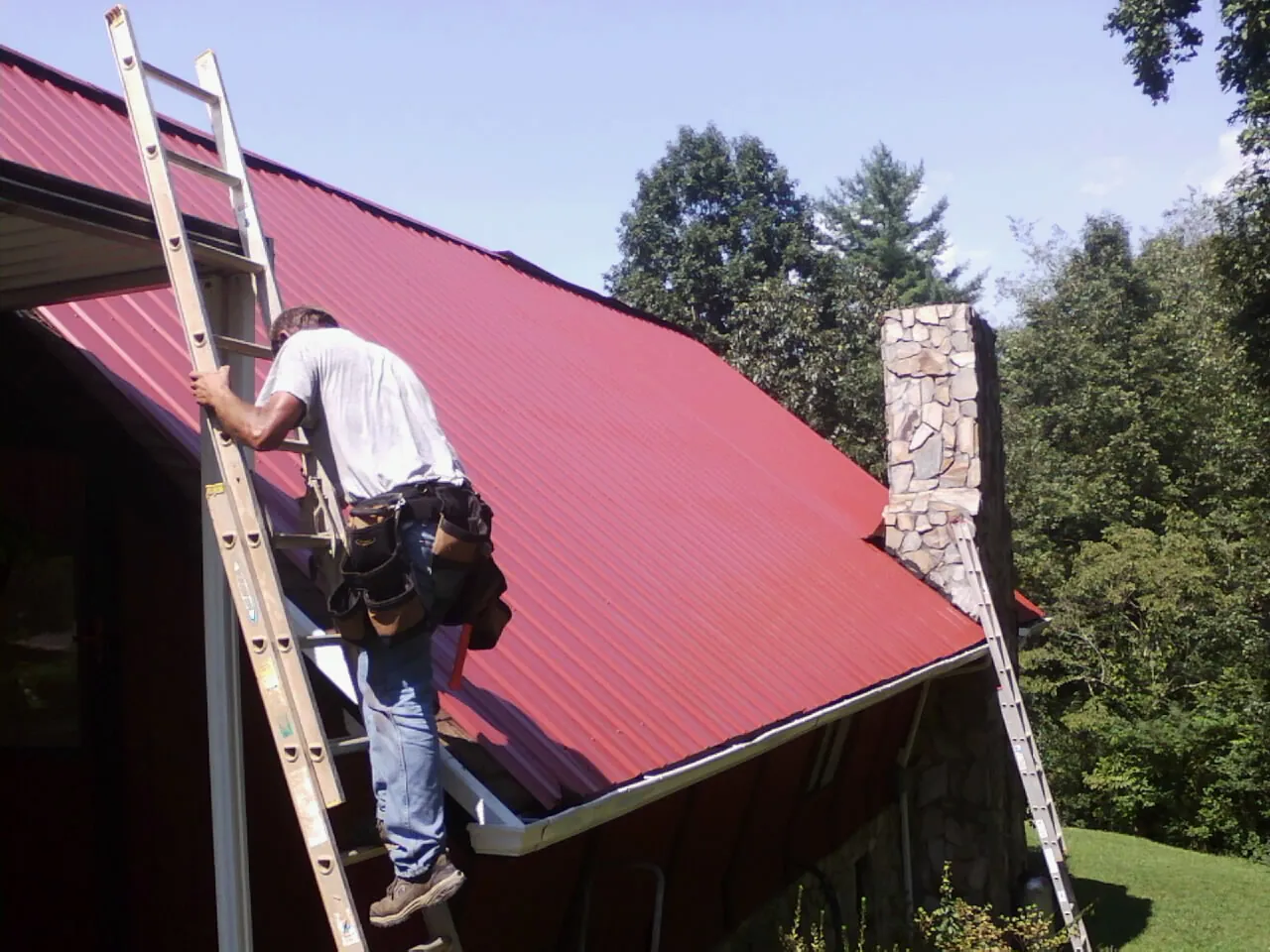 Roofer on a ladder beside a red metal roof and stone chimney — Soffit & Fascia Repair by Roofing Brothers in Statesville